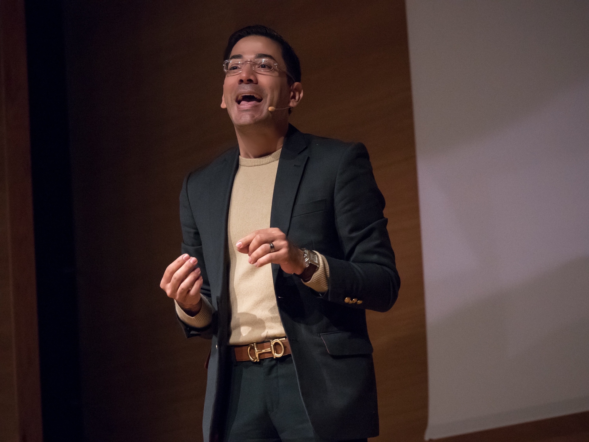 José Namén speaking on stage with a headset microphone, dark blazer and beige sweater, warm wooden backdrop.