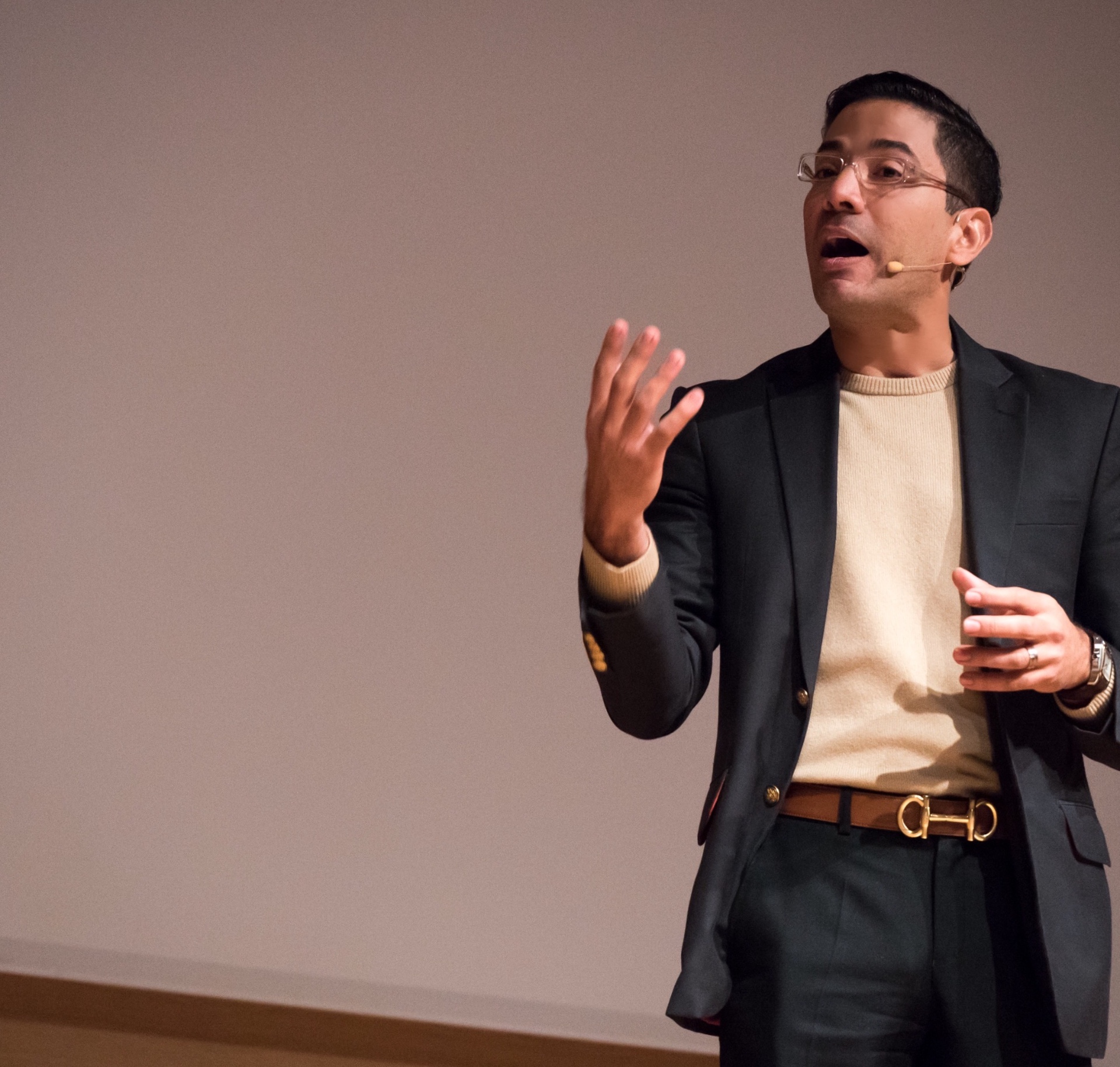 José Namén mid-talk on stage, raising one hand, wearing a headset microphone.